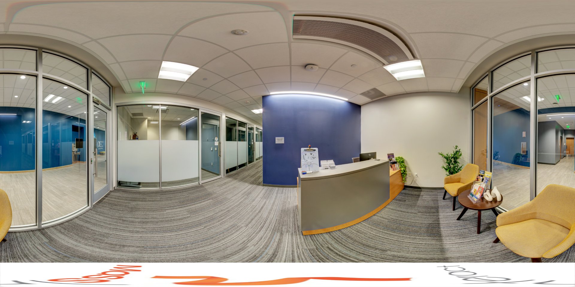 A panoramic view of a small office reception area with a curved desk, yellow chairs, and a small table. Glass walls separate the space from a hallway, and a blue accent wall adds color to the modern, clean design.